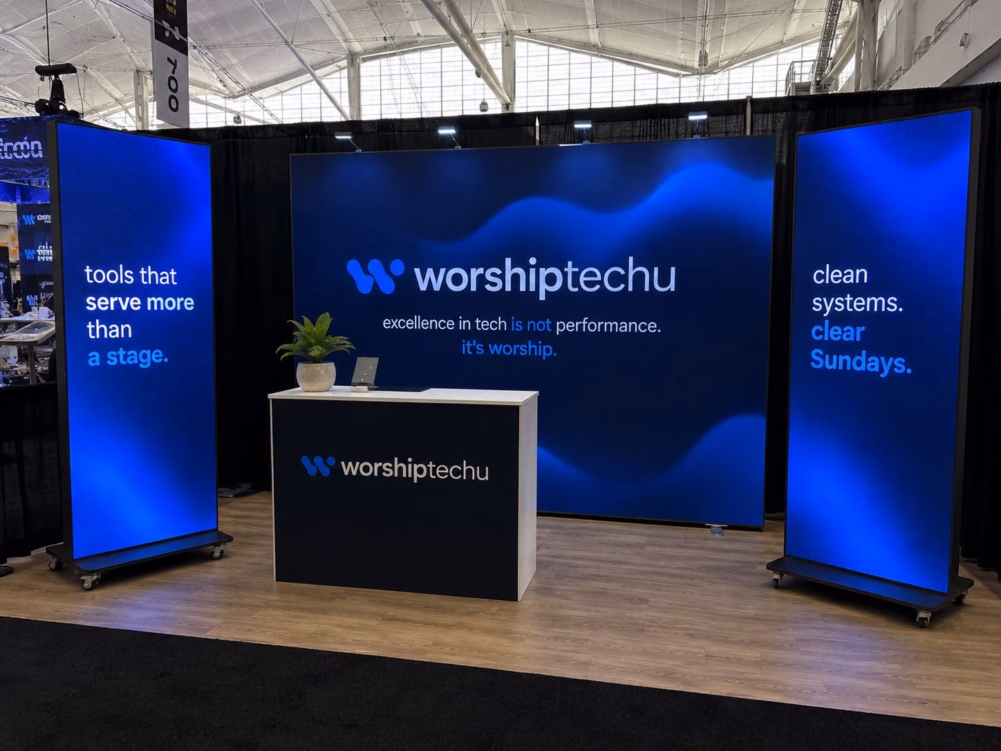 A trade show booth: a wide LED wall for worshiptechu between two LED totems, with a branded counter in front.